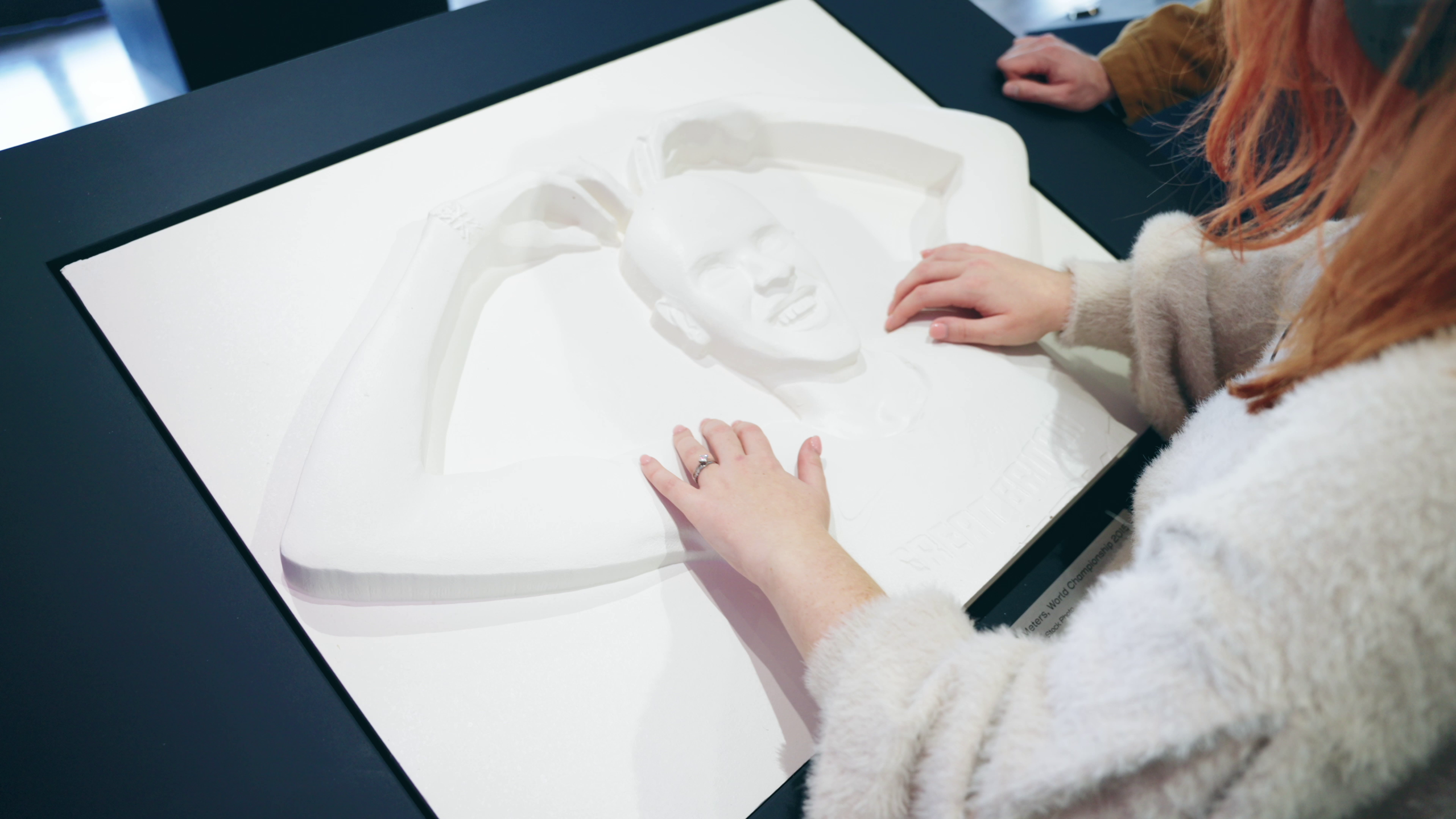 A young lady feels a tactile carving of Mo Farah and understands what he looks like for the first time. Mo Farah is standing in his classic 'Mobot' pose he made famous, with his arms in an 'M' shape above his head.